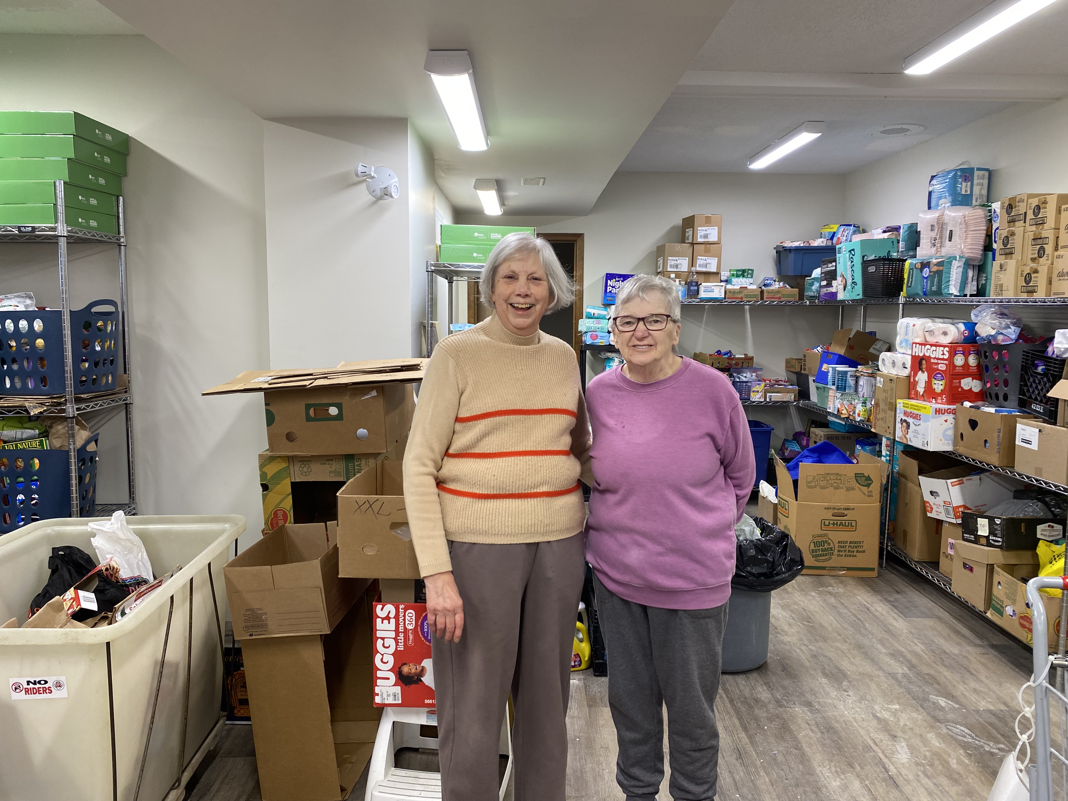 Two volunteers standing together in a food bank storage room, surrounded by shelves of donated food and stacks of boxes.