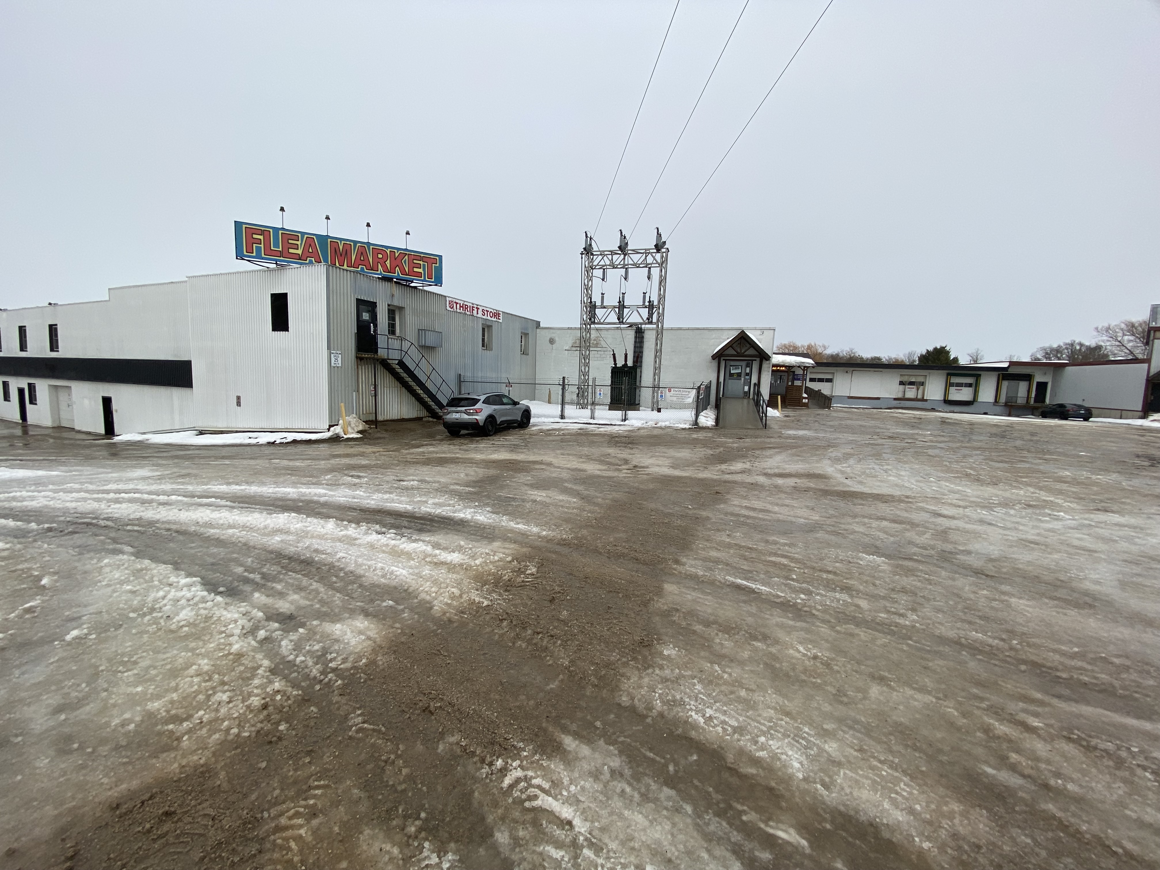 Wide view of a snowy parking lot in front of a large white building with a ‘Flea Market’ sign and external staircase.